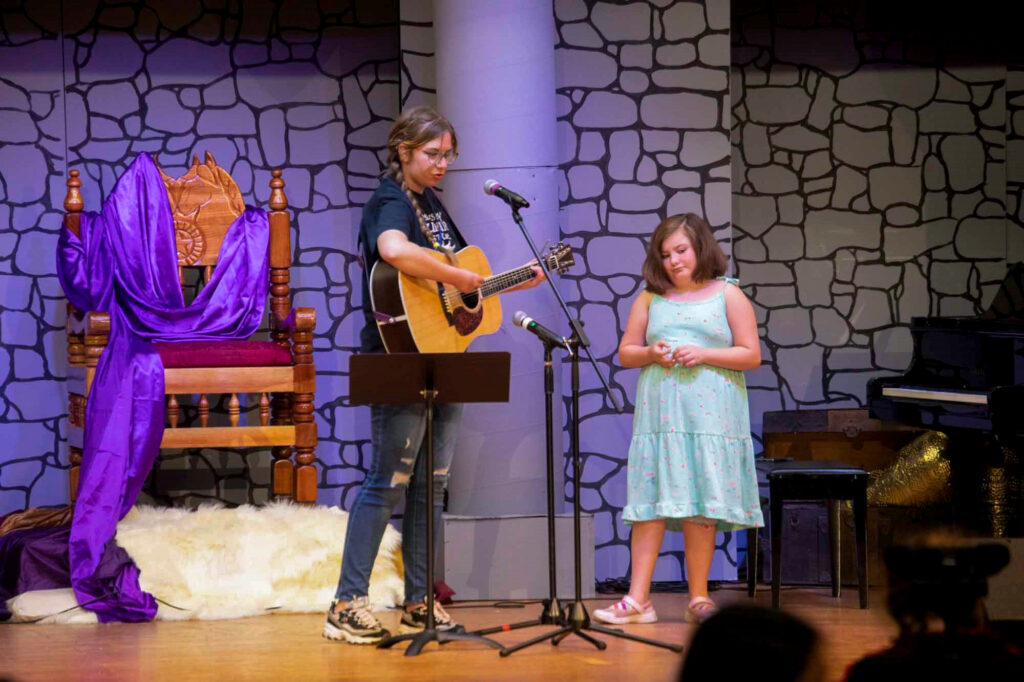 Evangeline and Cosette on stage for the talent show. Cosette is playing the guitar.
