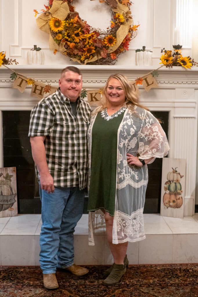 Maribeth and her husband smiling with their arms around each other in front of a fireplace decorated for Thanksgiving.