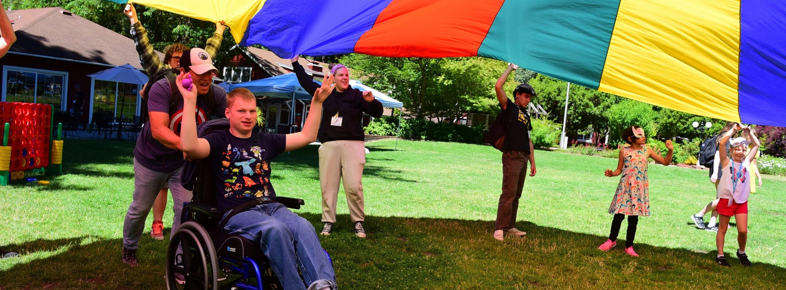 A young man in a wheelchair is being pushed under a large billowing parachute. His arms are raised and has a huge smile on his face.
