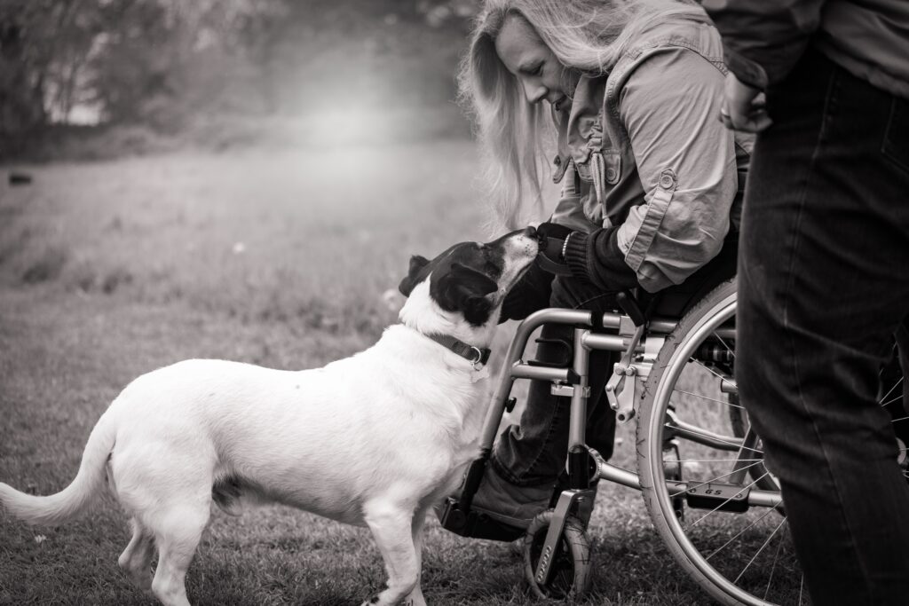A black and white photo of woman using a wheelchair looking down at a dog that has it's nose rested on her hand.