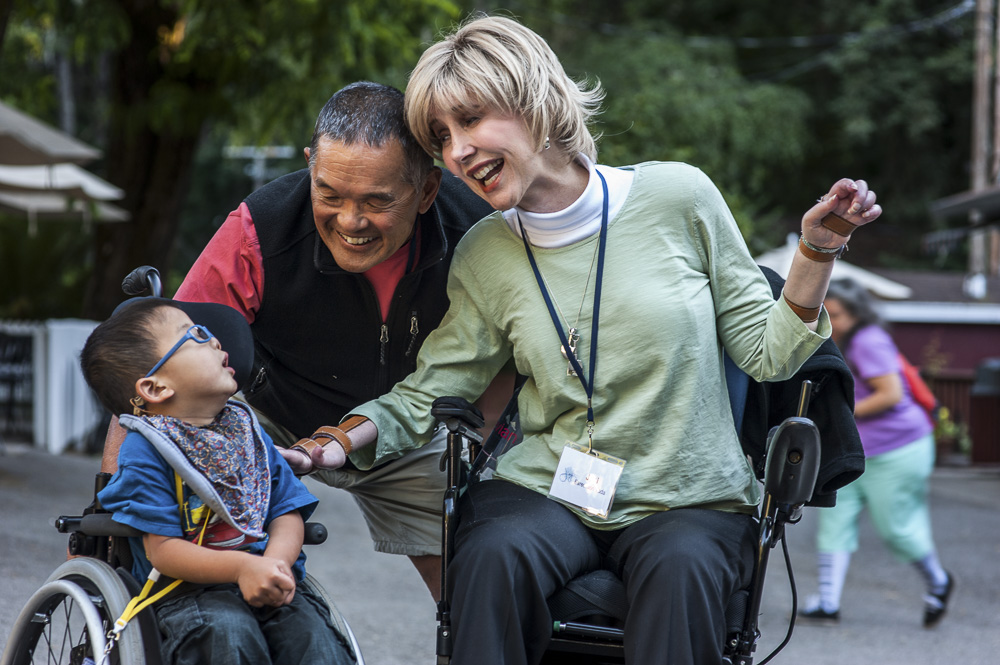 A picture of Joni seated in her wheelchair saying hi to a little boy seated in his wheelchair with Ken beside them leaning down and smiling.