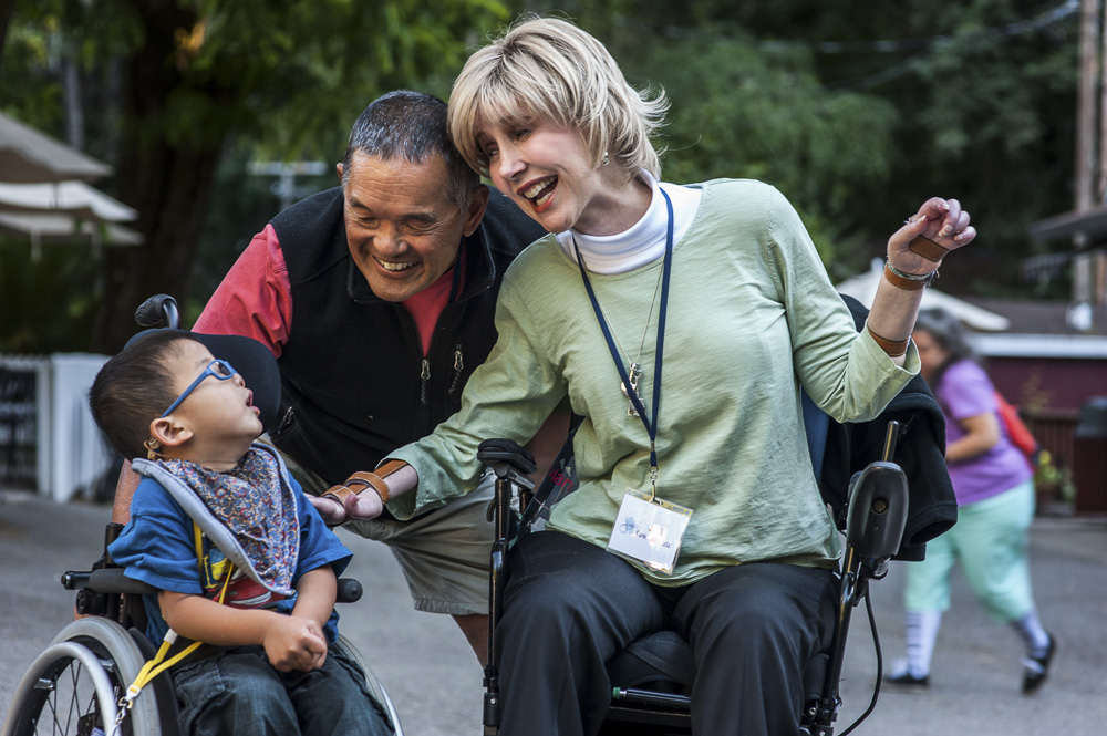 A picture of Joni seated in her wheelchair saying hi to a little boy seated in his wheelchair with Ken beside them leaning down and smiling. 