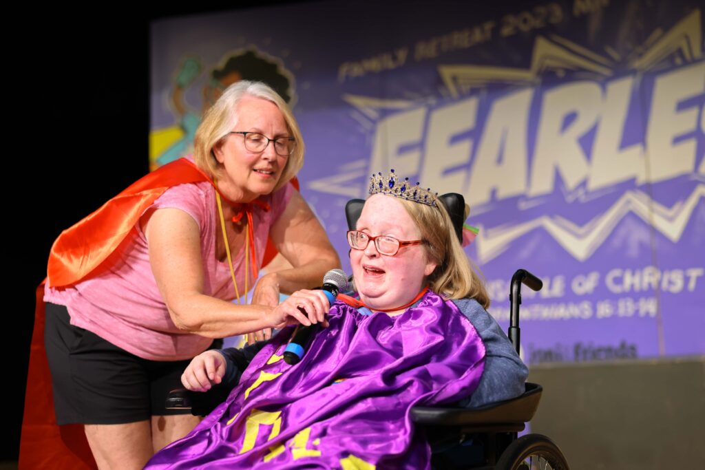 Jodi on stage as she's seated in her wheelchair with a crown on, her Joni and Friends buddy holding a mic up to Jodi's mouth as she shares her testimony.