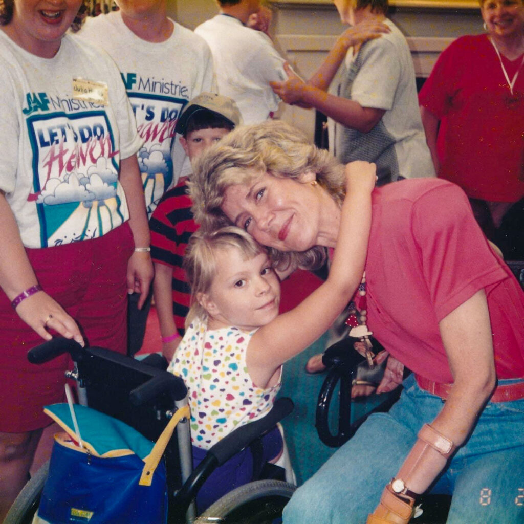 A picture of Jessica as a little girl giving Joni a hug around the neck as they're both seated in their wheelchairs and smiling at the camera.