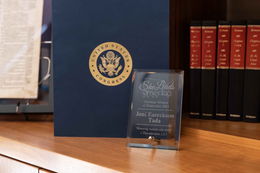 A picture of the She Leads America award on a wooden table with a dark blue folder behind it with a gold seal on it. The award itself is glass with words etched into it.