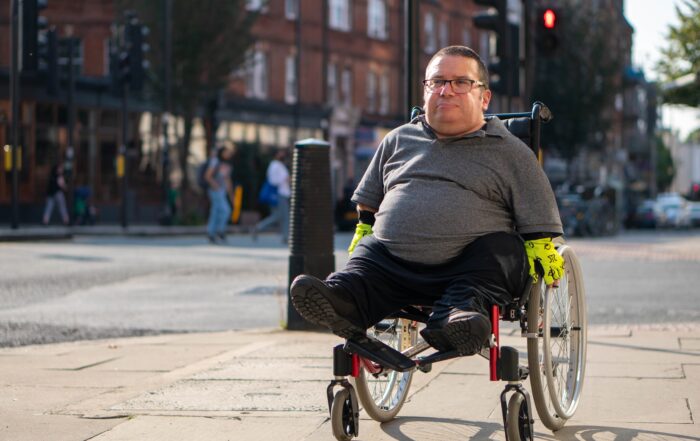 A man seated in a wheelchair on a street corner smiling.