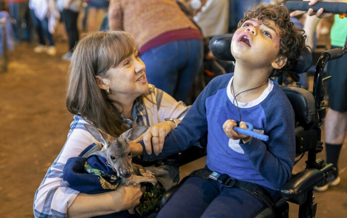 An older woman smiling as she holds a kangaroo in her arms. Nick is petting the kangaroo as he's seated in his wheelchair, looking up at sky.