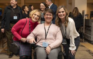 A woman seated in her wheelchair with two other woman beside her, all smiling at the camera.