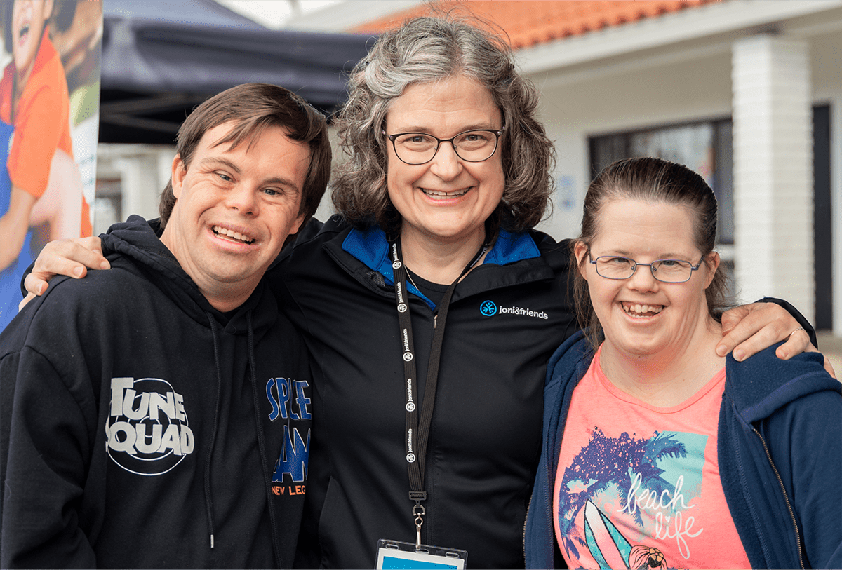 A picture of a Joni and Friends employee with her arms around two people with Down Syndrome as they all face the camera and smile.