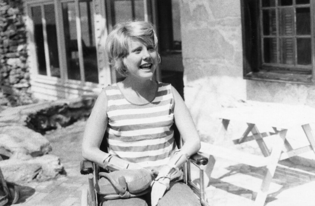 Young Joni sits in her wheelchair and is looking past the camera smiling.