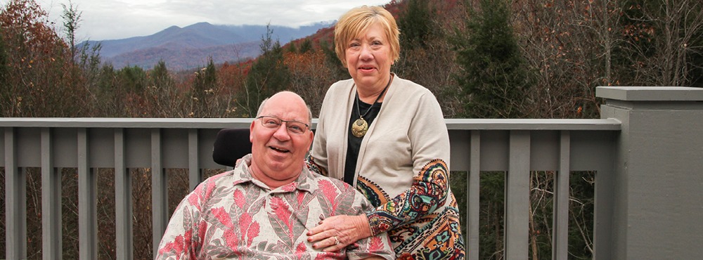 A smiling couple stand in front of a relaxing outdoor setting at Marriage Getaway. The husband is in a wheelchair.