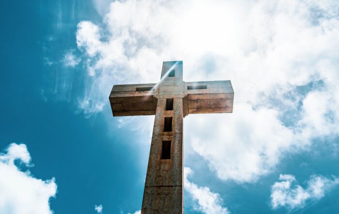 A picture from below a cement cross with clouds and blue sky above.