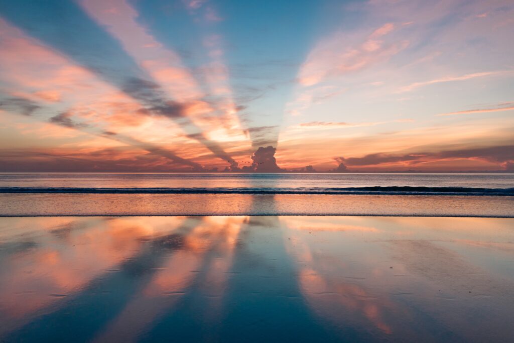 A picture of a seashore as the sun is setting on the horizon. The placement of the clouds is making the sun shine in beautiful rays.