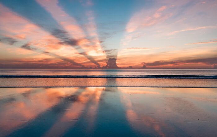 A picture of a seashore as the sun is setting on the horizon. The placement of the clouds is making the sun shine in beautiful rays.