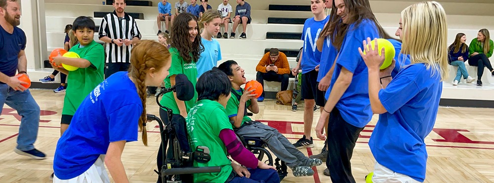 A group of smiling kids, some in wheelchairs, and teen volunteers participate at a dodgeball tournament.