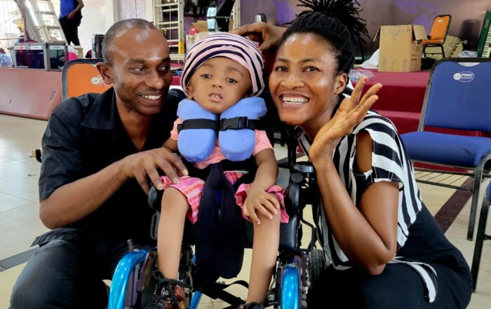 Melissa seated in a blue pediatric wheelchair and her father and mother on either side of her smiling at the camera.