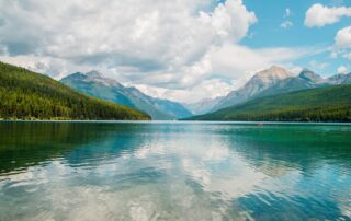 A picture of a crystal-clear lake with green hills and a mountain range surrounding it. Large white fluffy clouds are filling the blue sky.