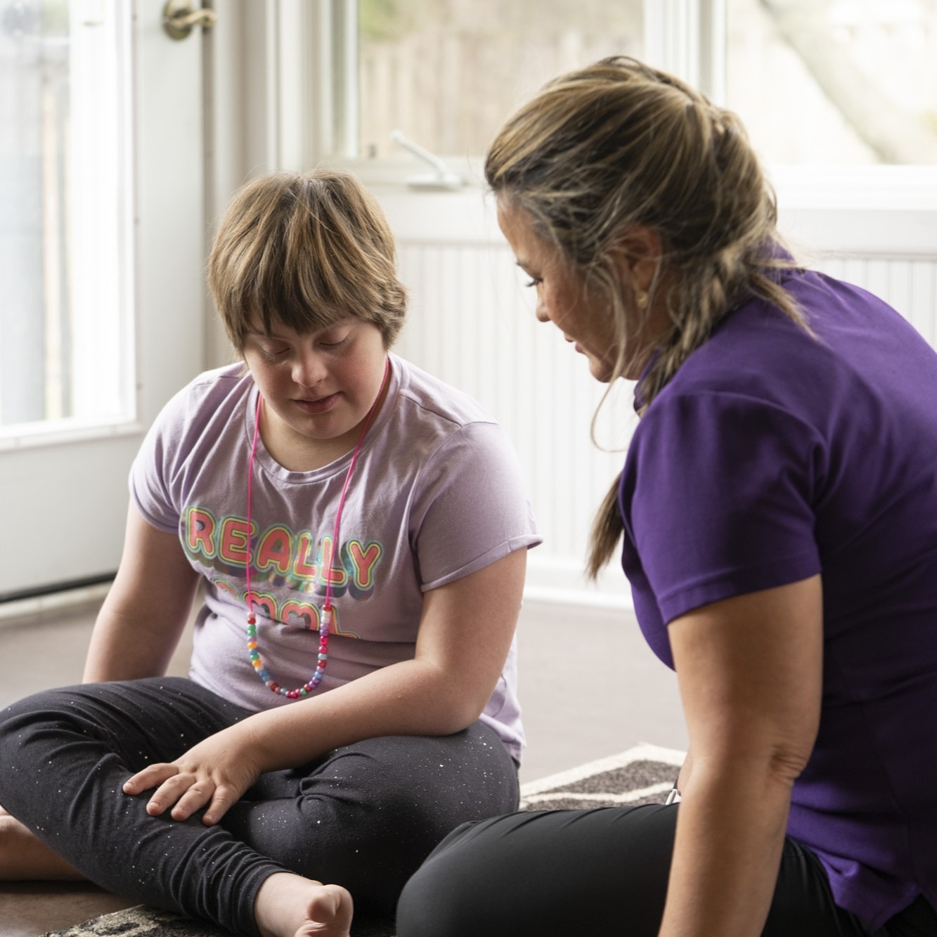 A young girl with Down syndrome sitting on the floor engaging in an activity with an adult woman.