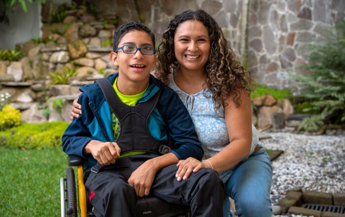 A picture of Ever in his new wheelchair with his mom beside him kneeling on the ground with her hand on his knee and the other hand on his shoulder. They're both smiling at the camera.