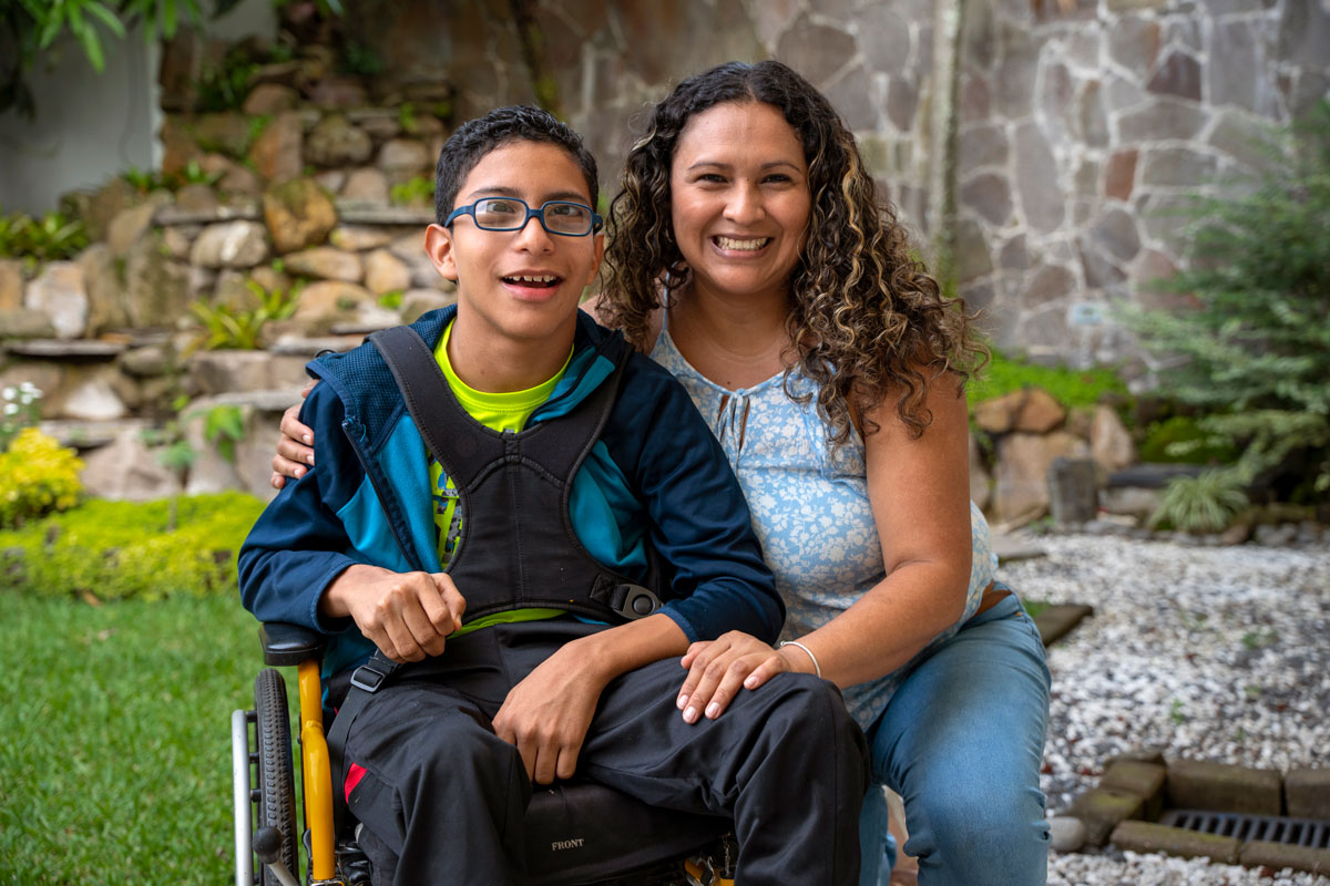 A picture of Ever in his new wheelchair with his mom beside him kneeling on the ground with her hand on his knee and the other hand on his shoulder. They're both smiling at the camera.