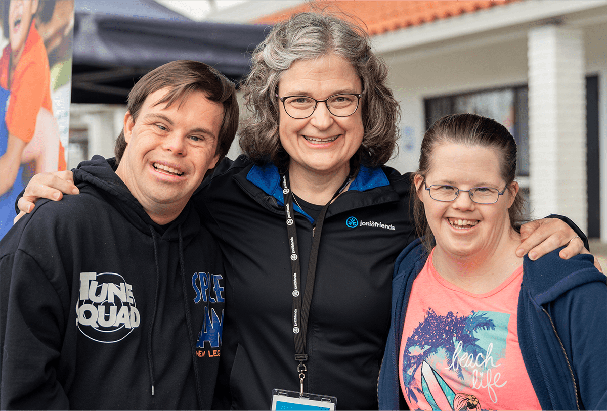 A picture of an older woman who is a Joni and Friends staff member with her arms around a young man with down syndrome on her left and a young woman with down syndrome on her right, all are smiling at the camera.