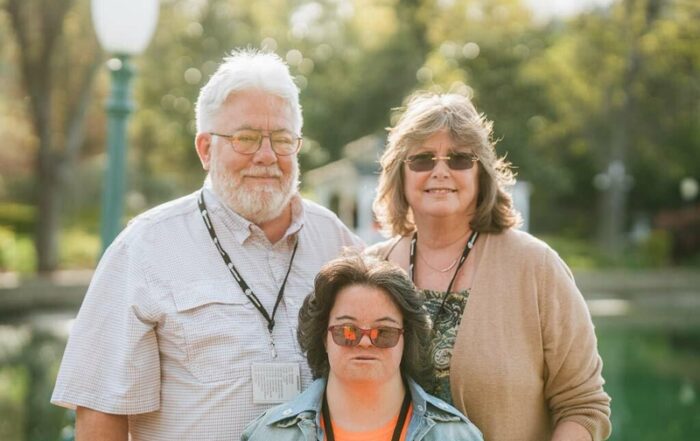 A photo of a mom, dad, and their daughter with down syndrome standing in front of a pond and smiling.