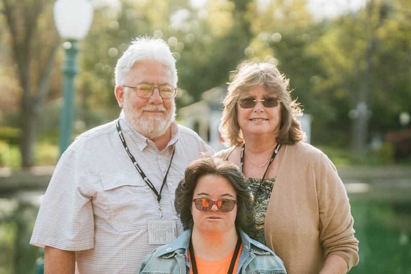 A photo of a mom, dad, and their daughter with down syndrome standing in front of a pond and smiling.