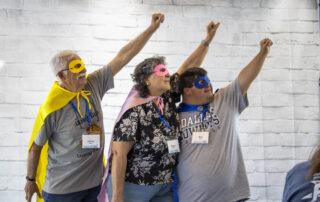 A picture of Jim, Nadine, and Ben in superhero masks and capes with their arms up in the air like they're going to fly.