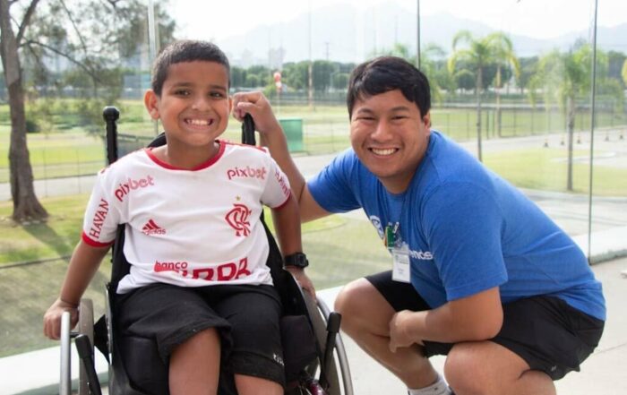 A picture of a young man with an intellectual disability serving as a Joni and Friends volunteer at an International Family Retreat. He's kneeling beside a young boy who is seated in a wheelchair.