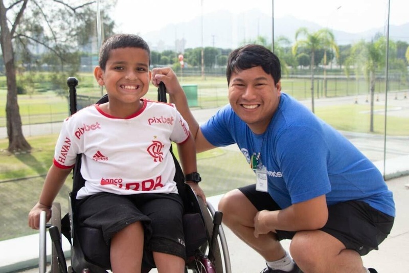 A picture of a young man with an intellectual disability serving as a Joni and Friends volunteer at an International Family Retreat. He's kneeling beside a young boy who is seated in a wheelchair.