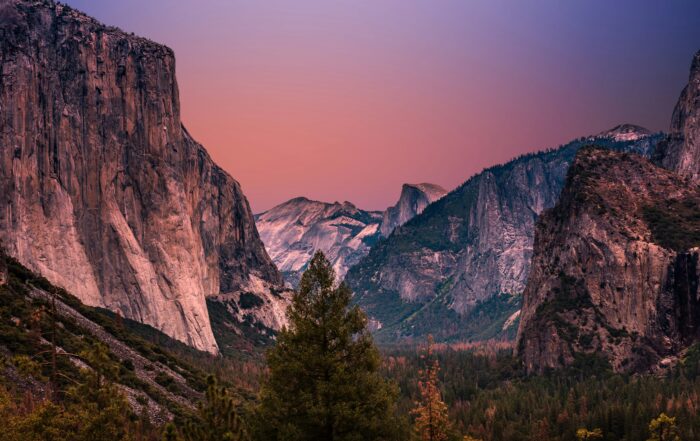 A picture of large, rugged cliffs with a beautiful, pink and blue sunset sky above. There is a forest of evergreen trees in the valley below.