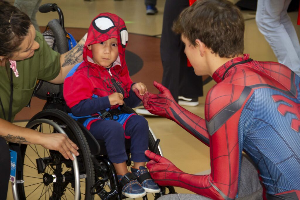 A picture of Jamie in a Spiderman costume with his Joni and Friends buddy also in a Spiderman outfit. Jamie is seated in his wheelchair and looking at the camera as he holds his Joni and Friends buddy's hand.