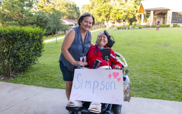 A picture of Jennifer seated in her wheelchair smiling at the camera with her mother Anne standing next to her.