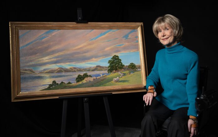 A picture of Joni sitting next to her Sermon on the Mount painting as she's seated in her wheelchair smiling at the camera.