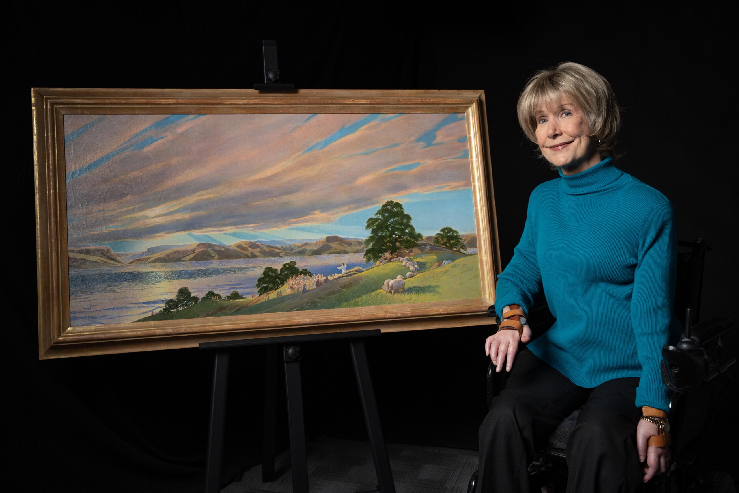 A picture of Joni sitting next to her Sermon on the Mount painting as she's seated in her wheelchair smiling at the camera.