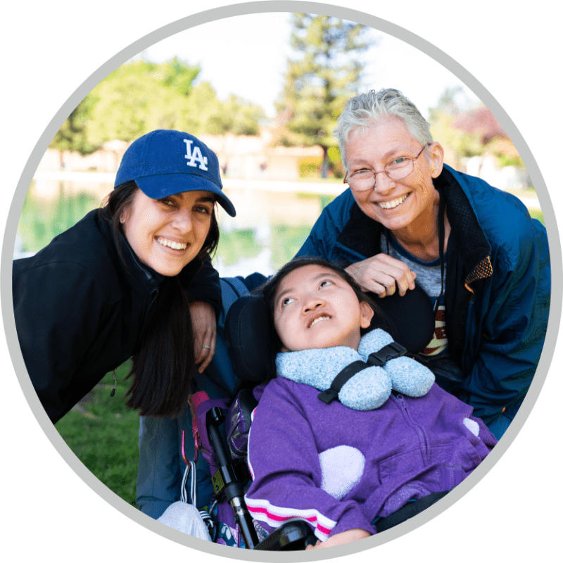 A girl in a wheelchair smiles with two women beside her, all three posing for the camera within a circular frame.