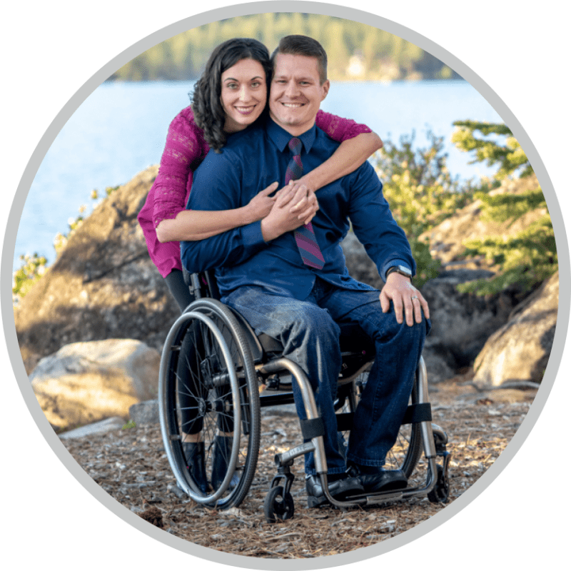 A woman hugging his husband who is in a wheelchair. Both are smiling and posing for the camera.