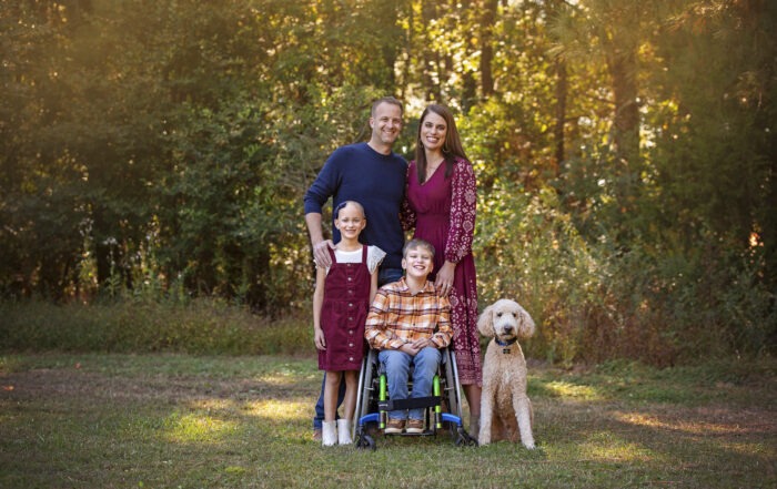 A group family photo of Karen, Chad, Claire, and Eli along with their dog. They are standing in front of a forest, surrounded by greenery.