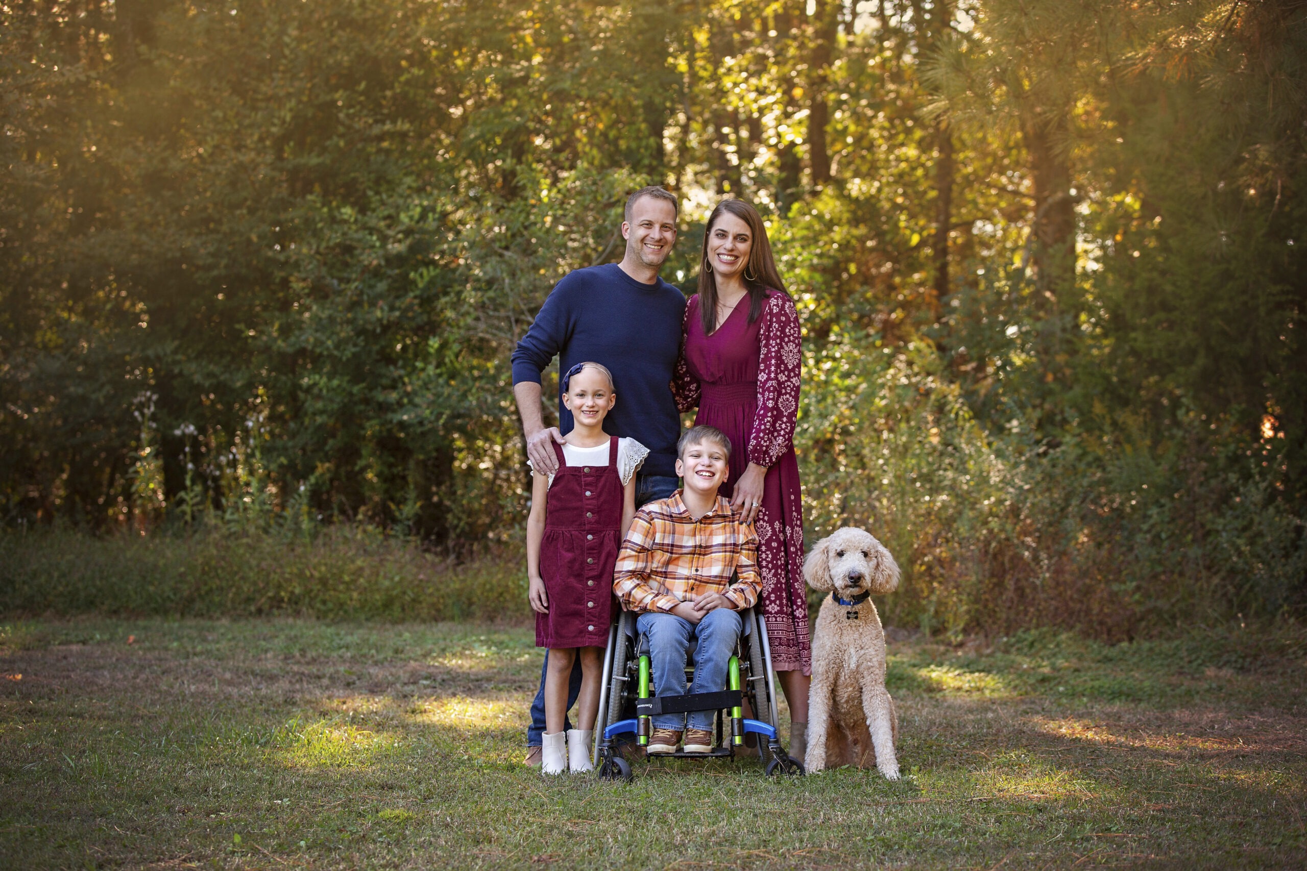 A group family photo of Karen, Chad, Claire, and Eli along with their dog. They are standing in front of a forest, surrounded by greenery.