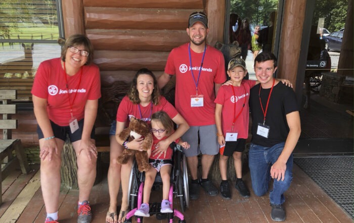 A group photo of Chris and Kayla at Knoxville Family Retreat with their children Morgan and Holsten. All are wearing their red Joni and Friends t-shirts. Little Morgan is seated in a small, pink detailed wheelchair.