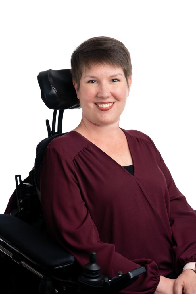 A portrait photo of Tracey seated in her wheelchair and smiling at the camera with a white background.
