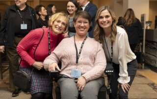 A picture of Tracy seated in her wheelchair smiling at the camera at a Joni and Friends all-staff conference with two other women on either side of her.