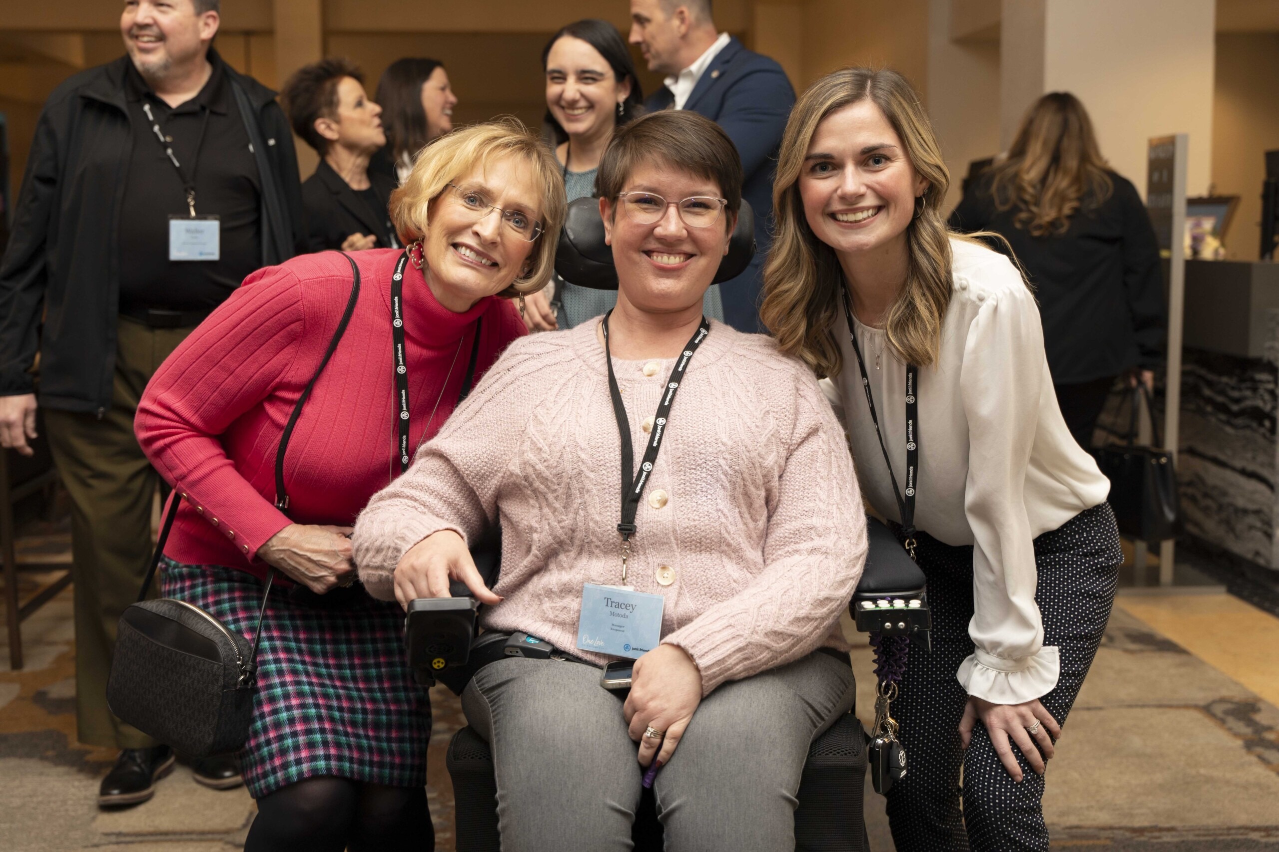 A picture of Tracy seated in her wheelchair smiling at the camera at a Joni and Friends all-staff conference with two other women on either side of her.