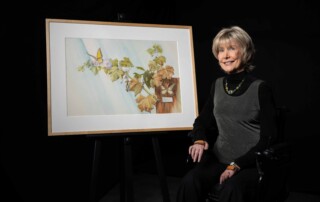 A picture of Joni sitting in her wheelchair next to her New Life painting and smiling at the camera.