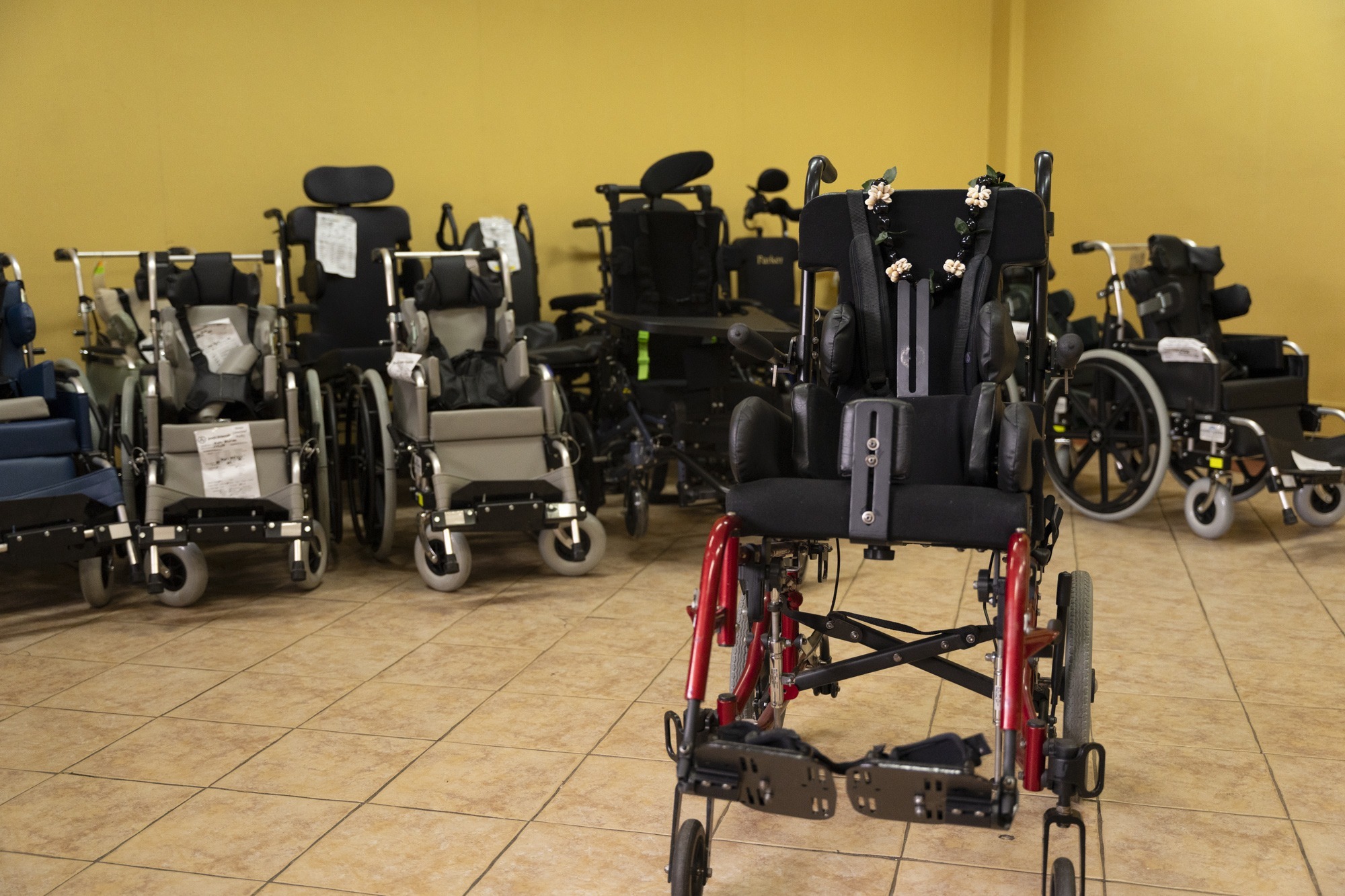 A picture of a small wheelchair with other wheelchairs lined up behind it against a bright yellow wall.