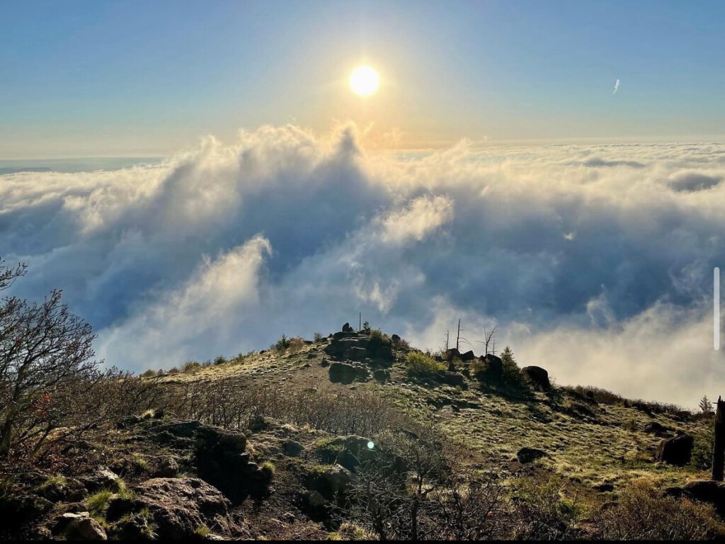 A view atop a mountain peak with clouds and the sun in the distance