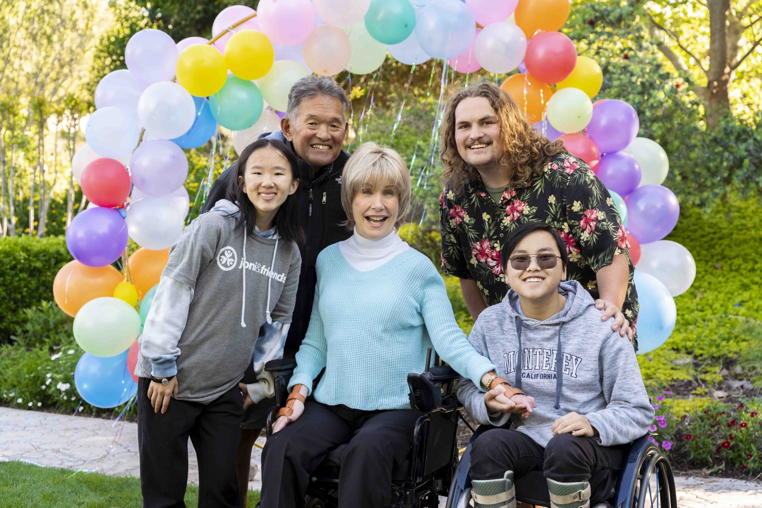 A group photo of Kylie seated in her wheelchair with Joni and Ken and two other people with her. All are smiling at the camera with a balloon arch behind them.