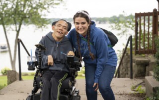 A picture of Da Wen seated in his wheelchair with his Joni and Friends volunteer buddy kneeling with her arm around him. Both are smiling at the camera with a beautiful body of water in the background.