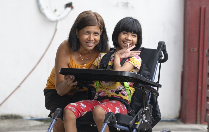 Chloe smiling as she's seated in her new wheelchair, her mother kneeling beside her.