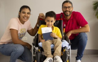 A group family photo of Patricia and Joel kneeling next to Matias who is seated in his new wheelchair. All are smiling at the camera. Matias is also holding his new Bible towards the camera with a big grin on his face.